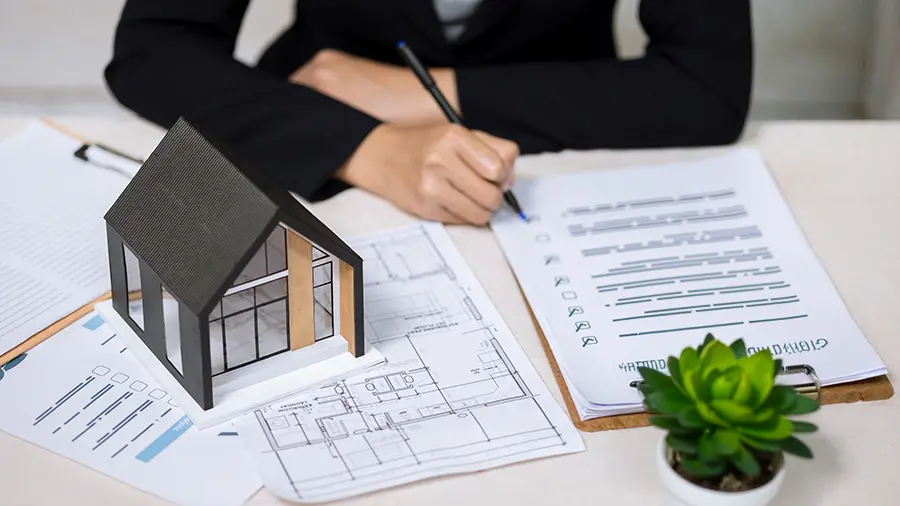 Businesswoman writing on document checklist near house model and architectural blueprints.